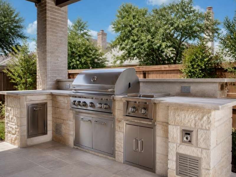 Light stone outdoor kitchen with built-in grill, ventilation openings, utility access panels, and electrical outlets in Fort Worth, Texas.